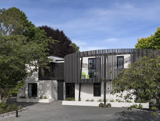 Exterior view of a contemporary multi-level residence set among mature trees. The white rendered facade is contrasted with dark vertical slatted screening wrapping around an upper balcony or facade element, with extensive glazing, a flat or very low-pitched roofline, and a gravel forecourt. The composition has clean geometric forms and strong screening elements integrated into the building envelope.