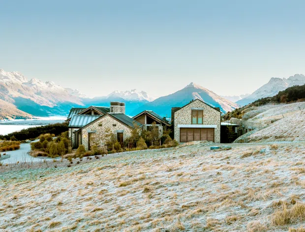 Exterior view of a low-rise stone-clad alpine house set on a sloping, frost-covered landscape beside a lake, with dramatic snow-capped mountain ranges in the background. The home has pitched gabled roofs, dark metal roof sections, timber-framed glazing, and a prominent integrated garage door.