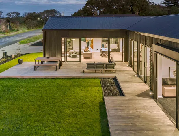 A low-profile contemporary house viewed from the landscaped exterior, with dark vertically clad walls and pitched metal roofs arranged around a timber-decked courtyard. Large glazed sliding doors open to a bright living area, while lawn, paving, and a gravel drainage strip frame the outdoor space.