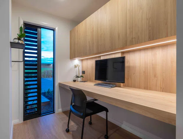 Compact, contemporary home office with a wall-to-wall timber built-in desk and overhead cabinetry. The joinery incorporates a recessed television or monitor, a slim integrated LED strip, and a black task chair. A tall glazed door or window with dark louvres provides natural light, while warm wood flooring and restrained decor create a clean, functional workspace.