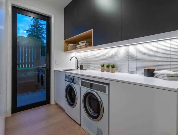 Contemporary compact laundry room with side-by-side front-loading washing machines beneath a long white benchtop. The room features dark matte overhead cabinetry, an open timber shelf, a white horizontally ridged tile splashback, a black-framed glazed exterior door, and warm timber-look flooring.