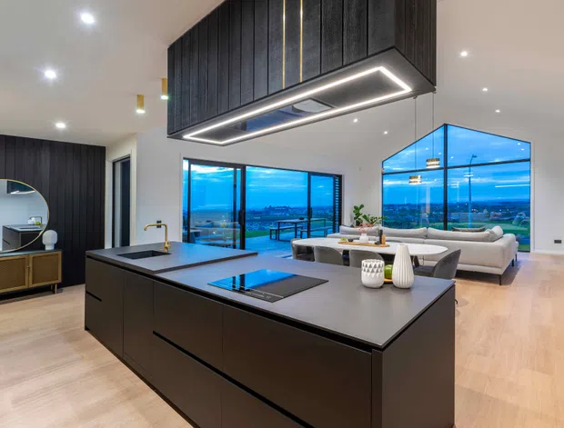 A spacious open-plan contemporary kitchen and living area with a large dark island, integrated cooktop, dramatic overhead rangehood enclosure, black vertical wall panelling, light timber flooring, and expansive blue-tinted glazing overlooking a distant landscape. A dining table and living seating are visible beyond the kitchen.