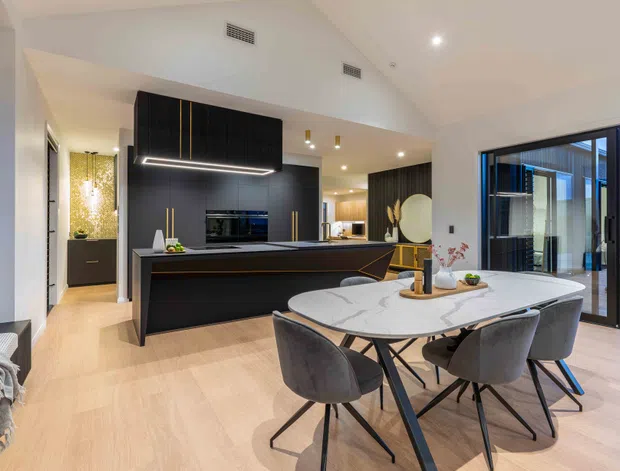Open-plan contemporary kitchen and dining area with a large black island, integrated dark cabinetry, pale timber flooring, and a round marble-look dining table with upholstered chairs. Floor-to-ceiling black-framed glazing opens toward an outdoor area, while warm recessed lighting and decorative accents create a refined, inviting interior.