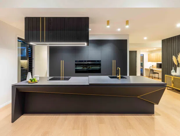 Contemporary open-plan kitchen dominated by a large dark island with a sculptural angular front, integrated black cabinetry, brass-gold accents and a suspended dark rangehood. Pale timber flooring, warm white walls, recessed lighting and a glimpse of an adjacent dining or living area create a refined, high-contrast interior.