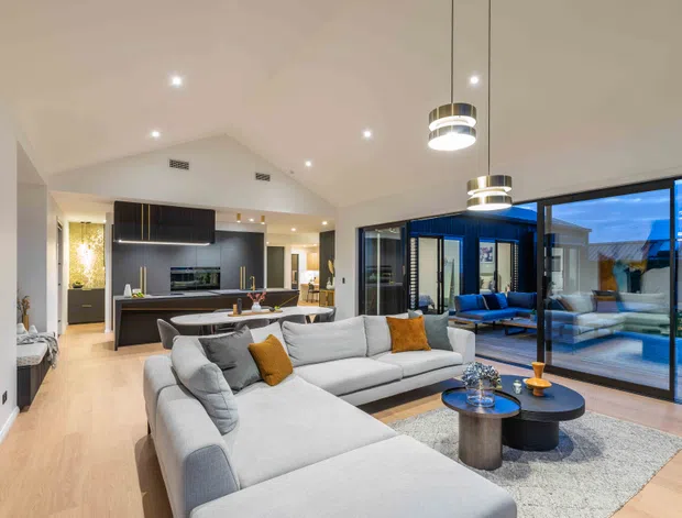 Contemporary open-plan living space with a large light-grey sectional sofa, round coffee tables, pendant lighting and a neutral textured rug. The living area connects directly to a sleek dark kitchen with an island, while expansive black-framed glazing opens toward an outdoor pool terrace. Light timber flooring, white walls and warm ambient lighting create an inviting, polished interior.
