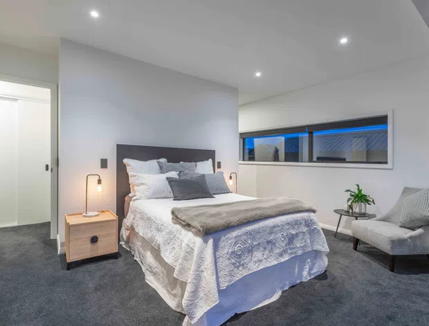 A contemporary bedroom with a neatly made bed, upholstered headboard, bedside table and lamp, grey carpet, recessed ceiling lights, built-in wardrobe doors, and a large horizontal window. The room uses a soft neutral palette with blue-grey bedding and a compact upholstered armchair.