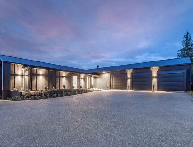 Twilight exterior view of a low, modern L-shaped residence or garage wing with dark metal cladding, extensive floor-to-ceiling glazing, warm interior lighting, and multiple wide garage doors. A broad paved driveway dominates the foreground, with restrained landscaping and a tall evergreen tree at the right.