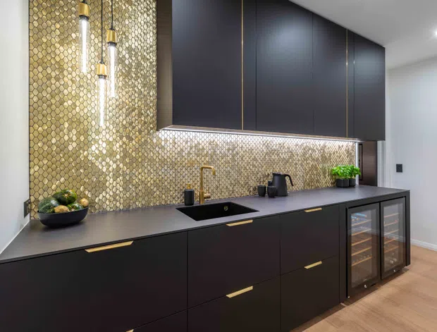 A sophisticated contemporary kitchen with matte charcoal cabinetry, slim brass pulls and accents, a dark benchtop, integrated black sink and brass tap. A gold-toned textured mosaic backsplash extends to the ceiling behind the cabinetry, complemented by pendant lights and under-cabinet lighting. Glazed beverage or wine-storage cabinets sit at the right, with warm timber flooring and minimal white walls.