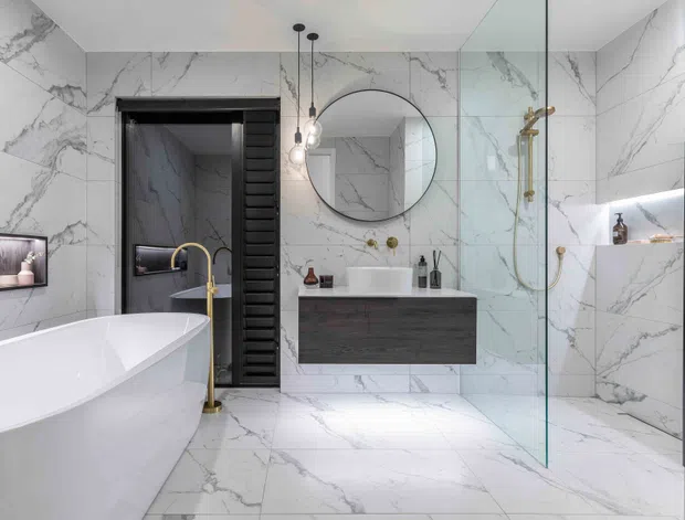 Luxurious contemporary bathroom clad almost entirely in white marble-look slabs with prominent grey veining. The room features a freestanding sculptural bathtub with a brushed brass floor-mounted tap, a floating dark timber vanity with a white vessel basin, a large round mirror, frameless glass shower enclosure, recessed shelving, black louvre-style door detailing, and a black heated towel rail. Pendant lights and brass fixtures add refined contrast.