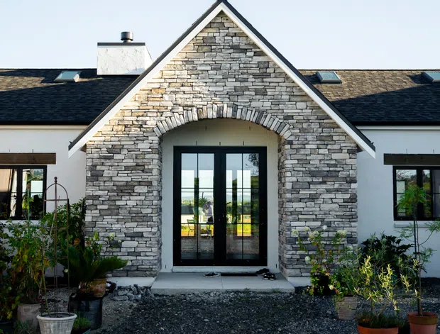 Front exterior view of a single-storey home with a prominent gabled entry porch clad in stacked grey stone veneer. The symmetrical facade has white rendered walls, dark roofing, black-framed glazed double doors, an arched stone surround, gravel landscaping, and potted plants.