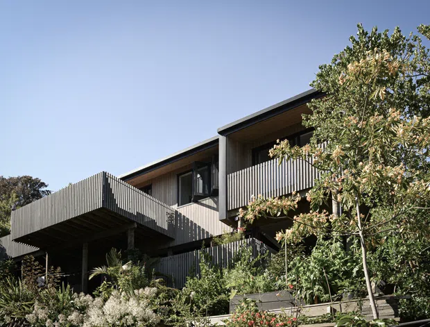 Exterior view of a contemporary multi-level residential home set within dense landscaped planting. The building features dark vertical ribbed cladding and screening, projecting balconies or decks, substantial cantilevered upper volumes, large windows, and a low-pitched roof with deep eaves.