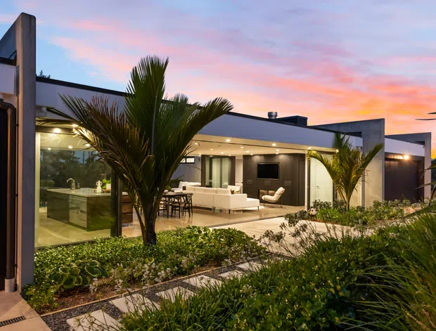 Contemporary single-storey home photographed at dusk, with strong geometric concrete or rendered walls, a broad flat roof, extensive floor-to-ceiling glazing, and a warmly illuminated open-plan kitchen and living area. The foreground contains landscaped garden beds with palms, ornamental grasses, and clipped shrubs, while a paved walkway leads toward the house. The pastel sunset sky forms the background.