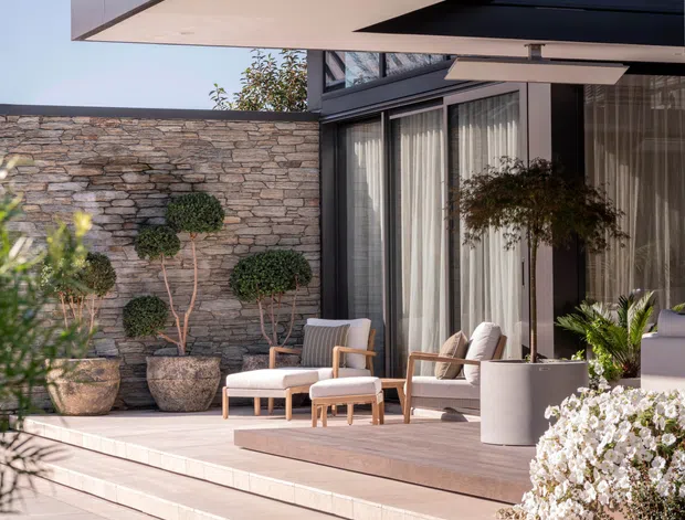 A contemporary outdoor terrace beside a modern home, featuring a broad cantilevered roof overhang, dark-framed floor-to-ceiling glazing, pale stone paving and stepped levels. The terrace is furnished with light upholstered lounge chairs and a low table, bordered by a stacked-stone privacy wall, sculpted topiary, potted plants and a manicured lawn.