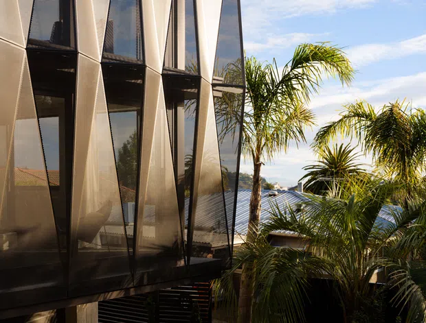 Exterior architectural view of a contemporary multi-level building with a strongly faceted facade composed of dark glazed vertical panels and angled metallic fins. Palm trees partially screen the building, with additional rooftops and blue sky visible in the background.