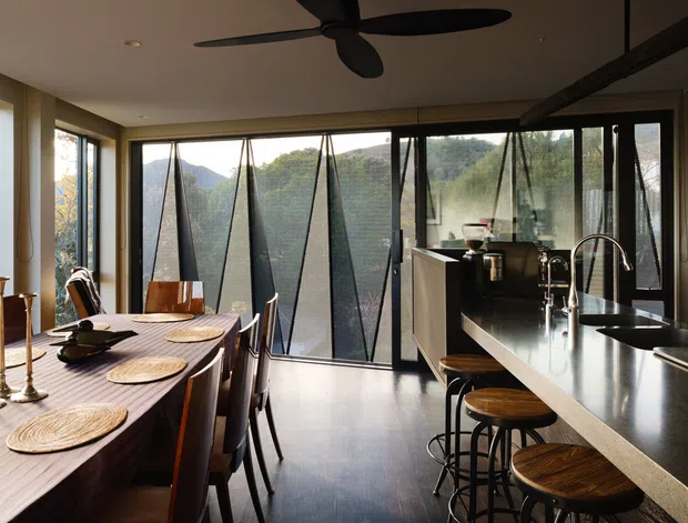 A contemporary open-plan kitchen and dining area with a long dark dining table, timber dining chairs, woven placemats, and decorative brass candlesticks. The kitchen features a large dark stone island with an integrated sink, curved faucet, black stools with timber seats, dark cabinetry, and extensive floor-to-ceiling glazing. Angled external sun-shading panels are visible through the windows, with leafy hills beyond. A large black ceiling fan and exposed dark beam add strong architectural character.