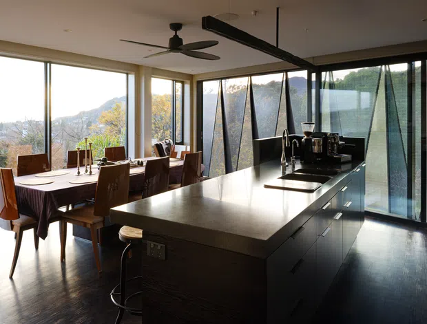 A modern open-plan kitchen and dining area with a large dark island, integrated cooktop and sink, dark cabinetry, and a timber dining table with upholstered chairs. Full-height glazing wraps the room, providing expansive views of a wooded, hilly landscape. A sculptural black ceiling fan and linear ceiling feature add contemporary detail.