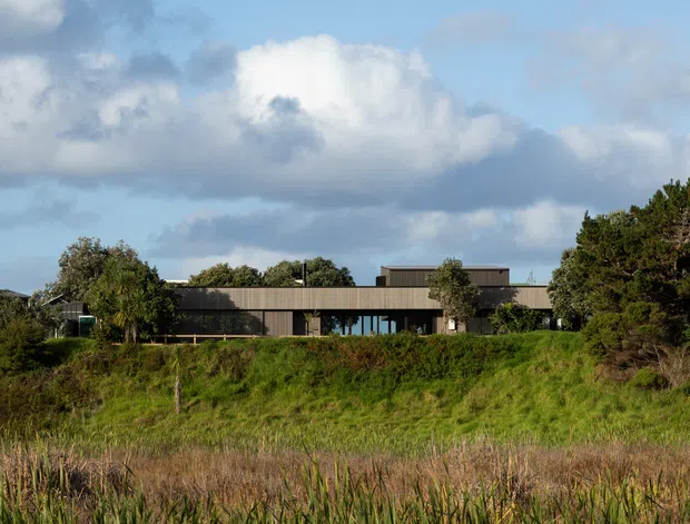 A low, horizontally composed modern house set within a coastal-looking grassy landscape. The building has dark vertical cladding, broad rooflines, extensive glazing, and a raised central volume, with dense vegetation surrounding the site. The image is an exterior architectural photograph under a partly cloudy sky.