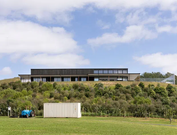 A wide exterior view of a long, low-slung contemporary house positioned on a gently elevated, landscaped site. The building has a dark horizontal upper volume, extensive glazing, projecting flat roof forms, and light-coloured lower walls. A broad lawn and dense native-style planting occupy the foreground beneath a partly cloudy sky.