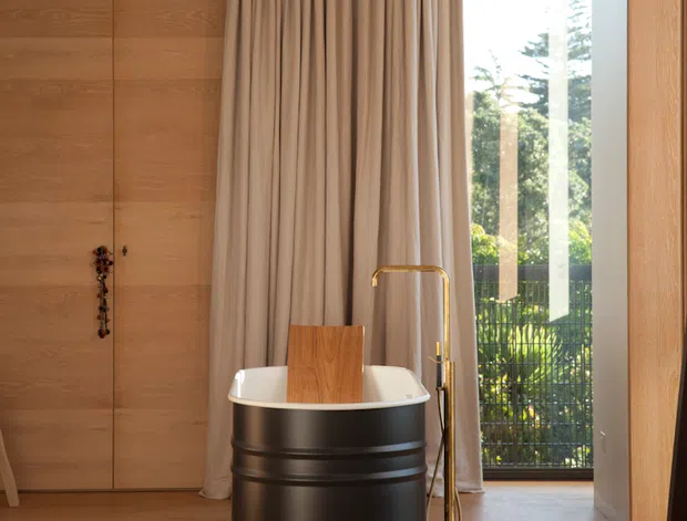 A warm, refined bathroom with a freestanding oval bathtub designed as a dark ribbed barrel-like form, paired with a brass floor-mounted tub filler. The room features extensive warm timber wall panelling and timber flooring, full-height taupe drapery, and a large glazed opening to greenery outside. The composition is minimalist and sculptural, with strong natural light and a calm material palette.