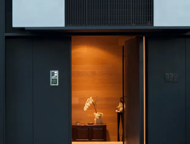 A contemporary residential entrance viewed from the street, featuring a dark charcoal facade and recessed entry door. The entry recess is lined with warm timber wall panelling and flooring, with a console cabinet, orchid arrangement, intercom, and house number 100. A tall upper-level opening is screened with fine vertical black metal bars. The foreground is paved in grey stone.