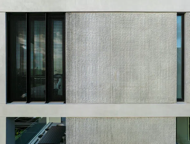 Exterior architectural view of a contemporary multi-storey facade. The composition features pale textured concrete or masonry panels, narrow black-framed vertical glazing with dark screening, and strong horizontal slab lines. A partial lower-level opening reveals an internal or covered circulation area. The facade is minimalist and rectilinear, with a cool neutral palette and high contrast between the light cladding and dark windows.
