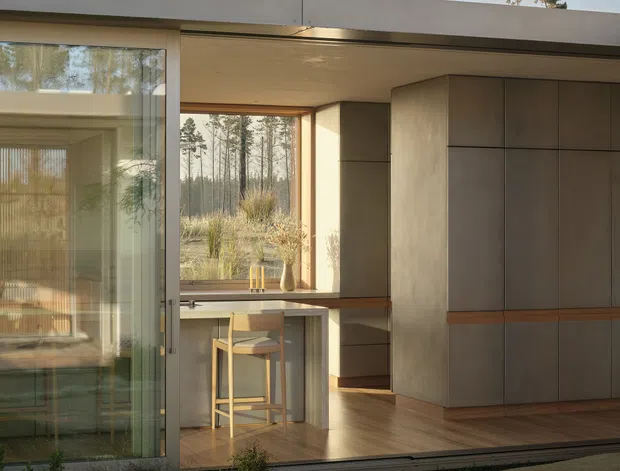 A contemporary single-storey flat-roof home viewed from an exterior courtyard or garden. Large glazed sliding openings reveal a warm, minimalist interior with timber flooring, a compact island or counter with stools, extensive built-in cabinetry, and a dining or living area. The architecture uses strong horizontal lines, floor-to-ceiling glazing, pale neutral surfaces, and integrated timber details, opening directly onto a landscaped outdoor area.
