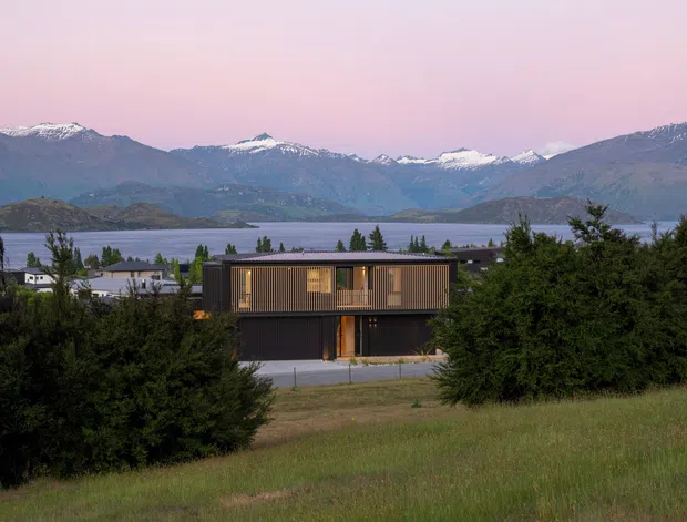 Exterior view of a contemporary two-storey rectangular house set among vegetation, with dark horizontal cladding, warm timber-toned vertical screening and a broad glazed upper level. The home overlooks a lake with snow-capped mountains beyond, viewed across a sloping grassy site.