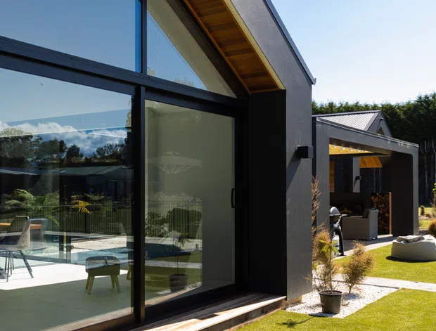 Contemporary residential exterior viewed from a landscaped garden, featuring dark rendered walls, expansive black-framed glazed doors and windows, a warm timber-lined eave, and a covered outdoor entertaining area. The glazing reveals a bright interior with dining and lounge furnishings, while paved paths, lawn, planting, and outdoor seating define the surrounding setting.