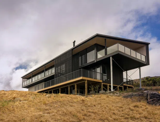 A contemporary multi-level house set on a sloping, sparsely vegetated site. The building has a dark linear facade with extensive glazing, projecting cantilevered upper volumes, recessed balconies, slim dark framing, and a low-pitched roof. The elevated construction responds to the terrain, with exposed supports beneath the main living levels. The image is an exterior architectural photograph under a partly cloudy sky.