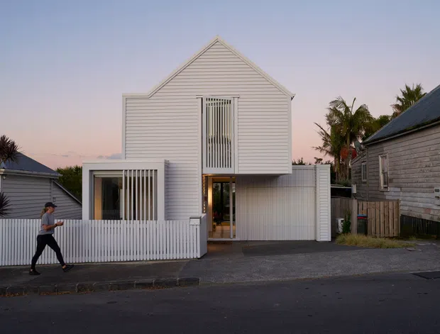 Street-level exterior view of a compact white weatherboard house with a steep central gable, tall vertical screened openings, a recessed entry, and a white picket fence. Neighbouring weatherboard homes, palms, and a paved driveway are visible in the residential streetscape at dusk.