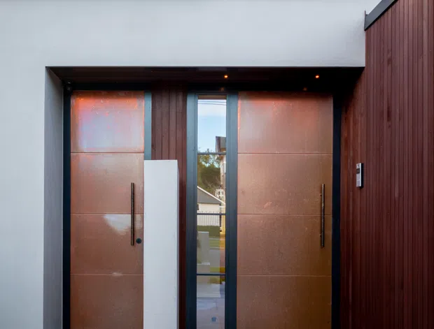 Contemporary residential entrance viewed from outside, featuring a recessed entry with large copper-toned metal doors, dark vertical timber cladding, pale rendered walls, grey paving, and a pitched gabled roof visible above. A narrow glazed panel beside the doors provides a view into the property.