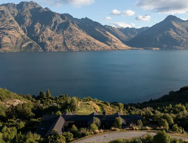 Aerial or elevated exterior view of a dark-roofed residential property set amid landscaped vegetation on a hillside, overlooking a broad deep-blue alpine lake surrounded by steep rugged mountains. The home has a low, modern roofline and blends into the sloping terrain.