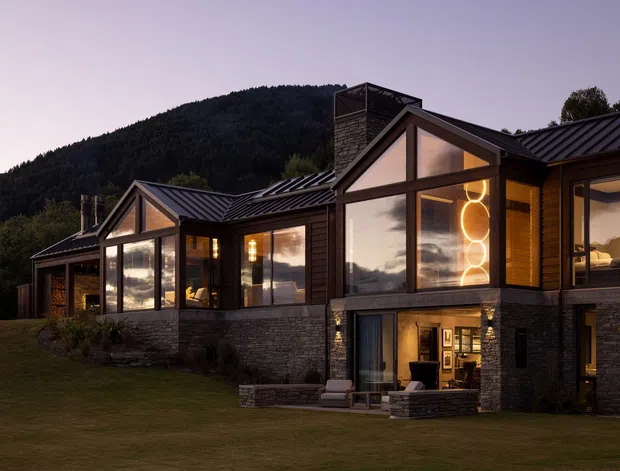 A large contemporary multi-level residence set against a forested mountain backdrop at dusk. The home features dark stone masonry, dark metal pitched roofs, extensive floor-to-ceiling glazing, timber-framed triangular gables, and warm interior lighting. A recessed lower-level outdoor sitting area and landscaped lawn are visible in front.