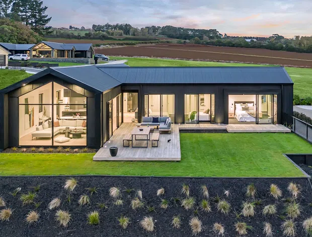 Aerial view of a contemporary single-storey home set within a landscaped rural site. The dark metal-clad house has a low pitched roof, extensive floor-to-ceiling glazing, a central timber deck with outdoor furniture, and a long rectangular swimming pool. Agricultural fields and neighboring rural buildings are visible beyond the manicured lawn.