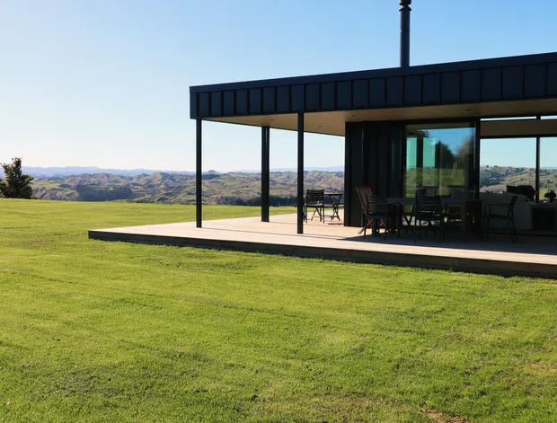 A low, modern pavilion-like house set in an expansive grassy landscape, with a flat roof, dark metal-clad fascia, slender black structural posts, extensive floor-to-ceiling glazing, and a broad covered outdoor terrace. The open-plan interior and outdoor dining area are visible through the glazing, with a dramatic rural or elevated view beyond.