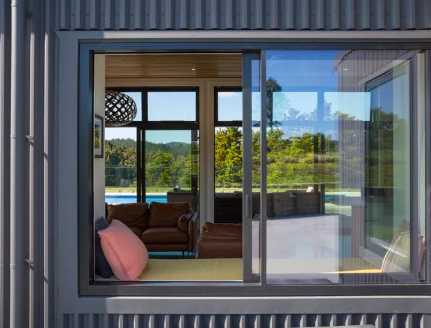 Exterior view of a contemporary home facade with dark grey vertical cladding and a large aluminium-framed sliding window. Through the glazing, a bright living room with a brown sofa, pink cushion, timber ceiling, and expansive views toward landscaped grounds and hills is visible. The image emphasises the window opening, facade materials, and indoor-outdoor connection.