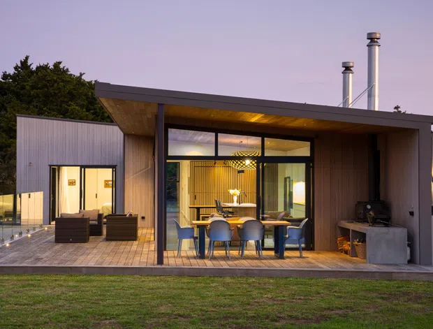 Contemporary single-storey pavilion-style home viewed from the lawn at dusk, with a low-pitched roof and deep timber-lined eaves. Floor-to-ceiling dark-framed glazing opens onto a timber deck containing an outdoor dining setting and lounge chairs. Warm interior lighting reveals a dining area and timber slatted feature wall, while an outdoor cooking or fireplace zone sits to the right. Vertical timber and metal-clad volumes extend to either side, with chimney flues rising above the roof.