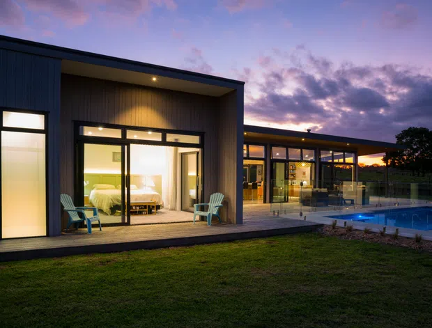 A modern single-storey residence photographed at dusk, with dark vertical cladding, broad floor-to-ceiling glazing, and a recessed bedroom terrace furnished with two outdoor chairs. The connected glazed living area extends toward a lit swimming pool, while warm interior lighting contrasts with the cool purple evening sky.