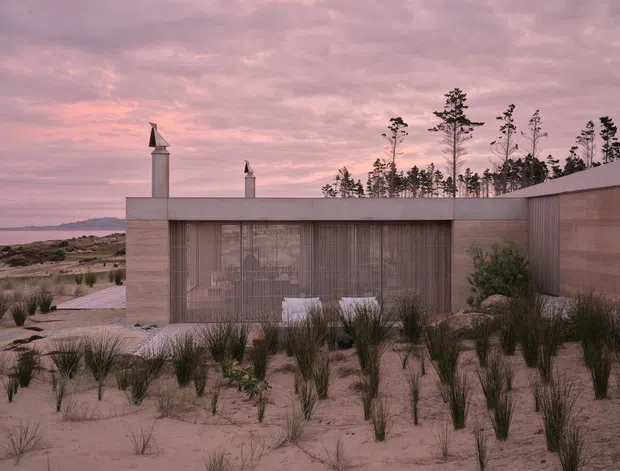 A low, contemporary coastal building set among sandy dunes and native grasses, viewed from the exterior at dusk. The rectilinear form has a flat parapet roof, pale rendered or concrete-like walls, expansive floor-to-ceiling glazing screened by sheer curtains, and a timber-toned side wall. Two slender chimney-like roof elements punctuate the silhouette. The muted pink sky, sparse coastal vegetation, distant shoreline, and restrained material palette create a quiet, atmospheric scene.