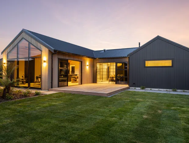 A contemporary single-storey home photographed at dusk from the landscaped front yard. The house has a dark grey metal-clad exterior, a pitched gabled roof, extensive floor-to-ceiling glazing, warm interior lighting, and a timber deck opening onto a lawn. The composition highlights the modern facade, glazed living spaces, and indoor-outdoor connection.