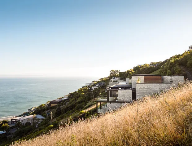 Wide exterior view of a modern residential development stepping down a steep, densely vegetated coastal hillside, with expansive ocean views and tall golden grasses in the foreground. The homes feature flat roofs, concrete-toned walls, dark glazing, and terraced forms integrated into the slope.