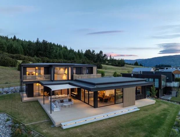 A contemporary multi-level residence set on a landscaped, sloping site with forested hills beyond. The home features dark exterior cladding, extensive floor-to-ceiling glazing, multiple low-pitched flat roof planes, balconies, and broad timber-toned outdoor terraces. Warm interior lighting glows through the windows, while a covered outdoor lounge and dining area extend from the main living spaces.