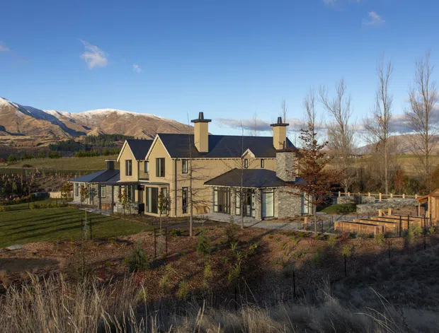 Exterior view of a substantial multi-gabled residence set in a landscaped rural or semi-rural valley. The house has pale rendered or masonry walls, dark pitched roofs, multiple chimneys, tall windows, covered verandah-like glazing, and timber fencing. Snow-dusted mountains and open cultivated grounds form the surrounding setting.