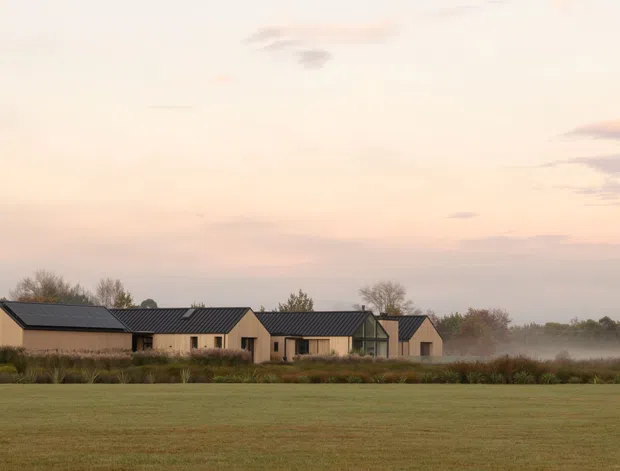 A low, single-storey rural residence viewed across open fields at dawn or dusk. The home has a long, clustered form with pale beige vertical cladding, dark pitched gabled roofs, and modest rectangular windows. Landscaping and misty vegetation surround the house, creating a quiet countryside setting.