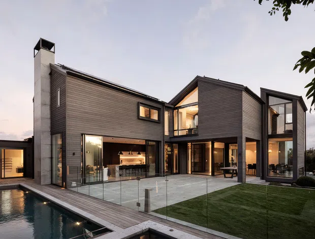 Contemporary multi-wing residence with dark horizontal cladding, extensive floor-to-ceiling glazing, and a flat-to-low-pitched roof composition. The house opens onto a paved courtyard and landscaped lawn beside a long narrow swimming pool, with warm interior lighting visible through the glass. A covered outdoor entertaining area and open-plan kitchen or living space are visible at ground level.