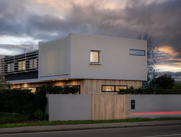 Contemporary two-storey residence viewed from the street at dusk, with a clean rectilinear form, pale rendered upper volume, vertical timber-like lower cladding, a projecting slatted balcony, narrow horizontal windows, and a solid front boundary wall with timber gate. Dark storm clouds and warm interior lighting create a dramatic evening atmosphere.