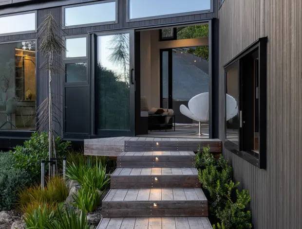 Exterior view of a contemporary multi-level home entry, showing dark vertical cladding, expansive black-framed glazing, a timber deck and stepped timber walkway with integrated step lights. The entry opens toward a refined interior with a sculptural white chair, while structured planting and stone landscaping frame the approach.