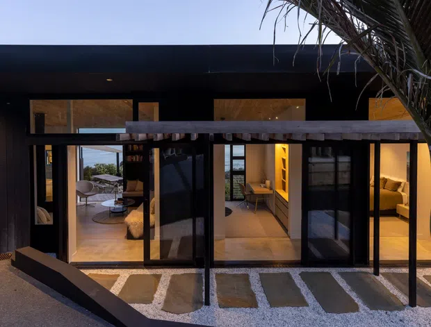 Twilight exterior view of a contemporary multi-level residence with a broad dark roof and extensive floor-to-ceiling glazing. Timber-toned interior spaces, furnishings, and warm lighting are visible through the glass, while a timber beam and palm fronds frame the foreground. A gravel-covered approach with rectangular stepping stones leads toward the entry.