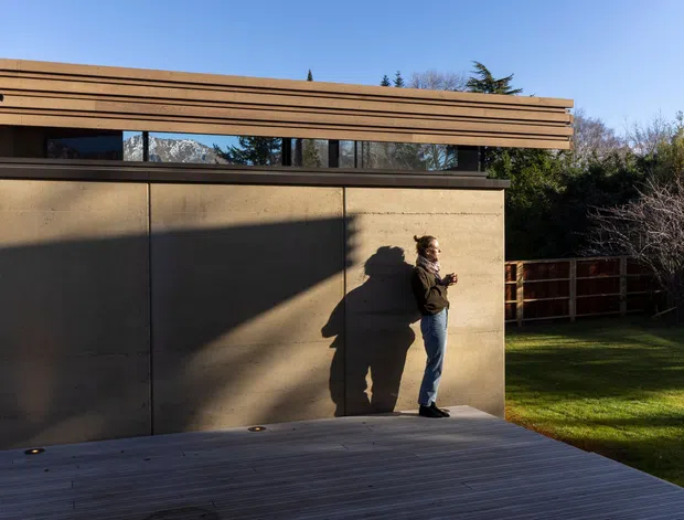 A contemporary single-storey architectural exterior viewed from a timber deck or covered outdoor area. The building features a long low horizontal form, smooth warm-toned rendered or concrete walls, narrow clerestory glazing, and layered horizontal timber fascia or screening. A person stands against the wall, with a fenced lawn and mature vegetation beyond. Strong afternoon shadows emphasize the minimalist geometric composition.