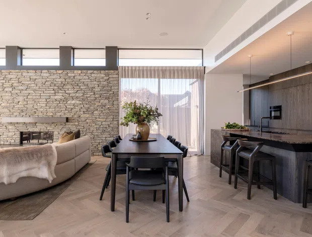 Open-plan contemporary living, dining, and kitchen area with a double-height feel, expansive clerestory glazing, a full-height stacked-stone fireplace wall, dark timber kitchen cabinetry, stone island benchtop, and light herringbone timber flooring. The room is furnished with a dining table, island stools, and a curved sectional sofa, creating a refined yet inviting interior.